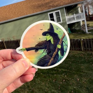 May include: A round sticker featuring a green-skinned witch in a black dress and hat, holding a staff. The background shows a sunset and silhouettes of birds. The witch is a character from the Wizard of Oz.