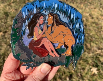 Può includere: Uno spillo in metallo argentato con un'immagine di cartone animato di un uomo e una donna in un ambiente di giungla. L'uomo è marrone e ha i capelli lunghi e marroni. La donna ha i capelli lunghi e marroni e indossa una maglietta gialla e una gonna rossa. Lo sfondo è una scena di giungla blu e verde.