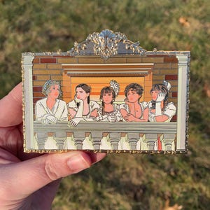May include: Gold-toned enamel pin depicting five women in white dresses standing on a balcony. The women are looking to the left and have different hairstyles. The pin has a detailed ornate frame.