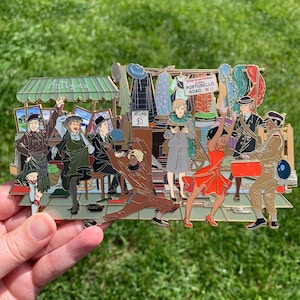 May include: A colorful enamel pin depicting a street scene with a market stall selling antiques. The stall has a green awning with the word "Antiques" written on it. There are several people in the scene, including a man in a kilt, a woman in a green dress, and a man in a brown suit. The pin is set against a green background.