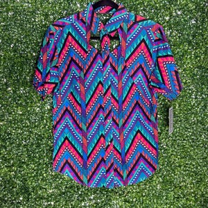 May include: Short-sleeved shirt with a vibrant geometric pattern in shades of blue, pink, teal, and black. The shirt is hanging on a black hanger against a green background. The design features chevron and dotted patterns.