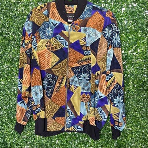 Könnte beinhalten: Eine farbenfrohe Bomberjacke mit einem geometrischen Muster in Blautönen, Braun, Gold und Lila. Die Jacke hat einen Reißverschluss und einen schwarzen gerippten Kragen und Ärmelbündchen.