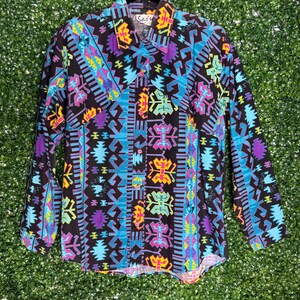 May include: A long-sleeved shirt with a colourful geometric design in black, blue, purple, yellow, and green. The shirt has a collared neckline and is suspended from a black hanger. The brand name "Sair" is visible on the collar.