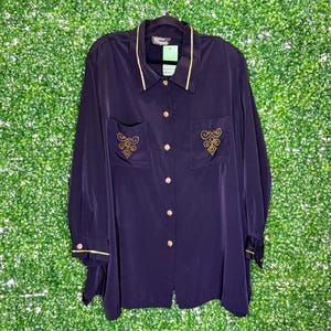 May include: A navy blue long-sleeved button-down shirt with gold trim and gold embroidered designs on the pockets. The shirt has a collar and a button closure.
