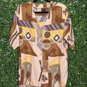 May include: A beige button-up shirt with a geometric pattern in brown, yellow, and purple. The shirt has a v-neck and short sleeves.