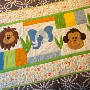 May include: A white quilted baby blanket with a green border and a row of colorful animal appliques. The animals include a lion, elephant, monkey, and zebra. The blanket is decorated with a floral pattern.