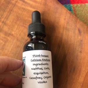 May include: A glass bottle with a dropper top filled with a dark liquid. The label reads "Plant-based Calcium tincture" and lists the ingredients: "Nettles, Oats, Equisetum, comfrey, organic Vodka".