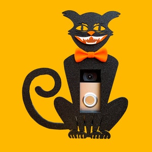 May include: Black cat silhouette with a bright orange bow tie and a doorbell camera mounted in the center of its chest. The cat is smiling with sharp teeth.