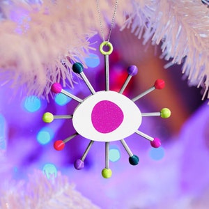 Puede incluir: Un adorno navideño de color blanco y rosa en forma de estrella con un centro rosa brillante y radios de metal plateado. El adorno está decorado con cuentas de colores.