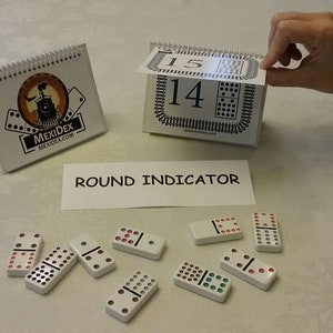 Könnte beinhalten: Ein weißes Tischspiel mit Dominosteinen und einer Rundenanzeigekarte. Das Spiel enthält eine Rundenanzeigekarte mit dem Text "ROUND INDICATOR" und eine weiße Karte mit der Nummer "14" und einem Dominomuster. Auf dem Tisch liegen außerdem mehrere Dominosteine, darunter ein Doppel-Sechs-Domino, ein Doppel-Fünf-Domino, ein Doppel-Vier-Domino, ein Doppel-Drei-Domino, ein Doppel-Zwei-Domino, ein Doppel-Eins-Domino und ein leeres Domino.