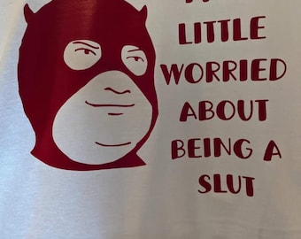 May include: White t-shirt featuring a red graphic of a masked face with the text "I'M A LITTLE WORRIED ABOUT BEING A SLUT". The graphic is a bold, simple design, and the text is in a sans-serif font.