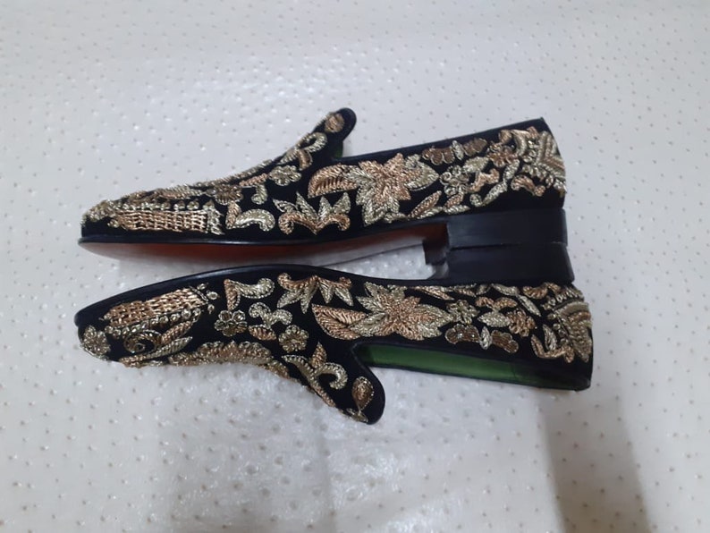 Puede incluir: Un par de mocasines de terciopelo negro con bordados dorados. Los mocasines tienen una punta puntiaguda y un tac&oacute;n bajo.
