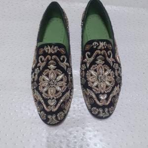 Puede incluir: Un par de mocasines de terciopelo negro con bordados dorados y plateados. Los mocasines tienen un forro verde y una punta puntiaguda.