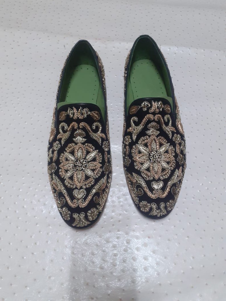 Puede incluir: Un par de mocasines de terciopelo negro con bordados dorados y plateados. Los mocasines tienen un forro verde y una punta puntiaguda.