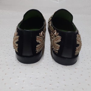 Puede incluir: Un par de mocasines de terciopelo negro con bordados dorados y plateados. Los mocasines tienen una punta puntiaguda y un tac&oacute;n bajo.