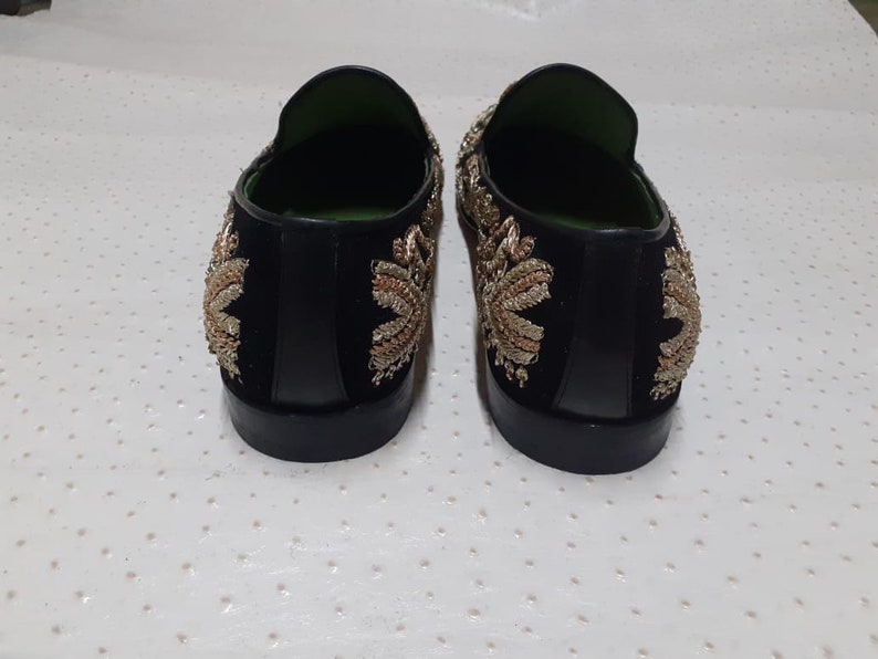 Puede incluir: Un par de mocasines de terciopelo negro con bordados dorados y plateados. Los mocasines tienen una punta puntiaguda y un tac&oacute;n bajo.