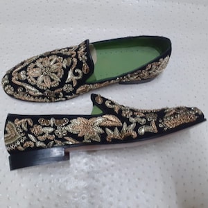 Puede incluir: Un par de mocasines de terciopelo negro con bordados dorados y plateados. Los mocasines tienen una punta puntiaguda y un tac&oacute;n plano.