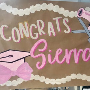 May include: A hand-painted sign on brown paper with the words "Congrats Sierra" in pink and white lettering. The sign includes a pink graduation cap, a pink bow, a pink hairdryer, and scissors. The border is white.