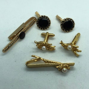 May include: A collection of gold-toned men's accessories, including tie clips, cufflinks, and tie tacks. The tie clip features a dark red stone, while the cufflinks have black stones and rope detailing. Some pieces have pearl accents.