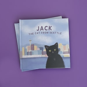 May include: Two children's books stacked on a purple surface. The top book cover features a black cat with yellow eyes, the text "JACK THE CAT FROM SEATTLE", and a Seattle skyline illustration.