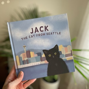 May include: A children's book titled "Jack the Cat from Seattle" featuring a black cat illustration with the Seattle skyline in the background. The book cover is light blue with white clouds and the title in large, bold letters.