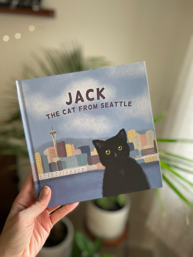 May include: A children's book titled "Jack the Cat from Seattle" featuring a black cat illustration with the Seattle skyline in the background. The book cover is light blue with white clouds and the title in large, bold letters.