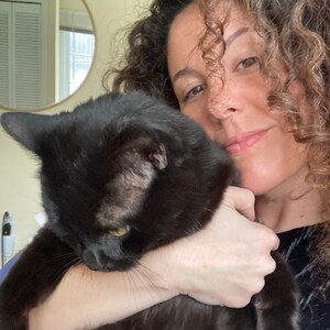 May include: A black cat with yellow eyes is being held. The cat's fur is shiny and smooth. The background includes a mirror and a person with curly hair.