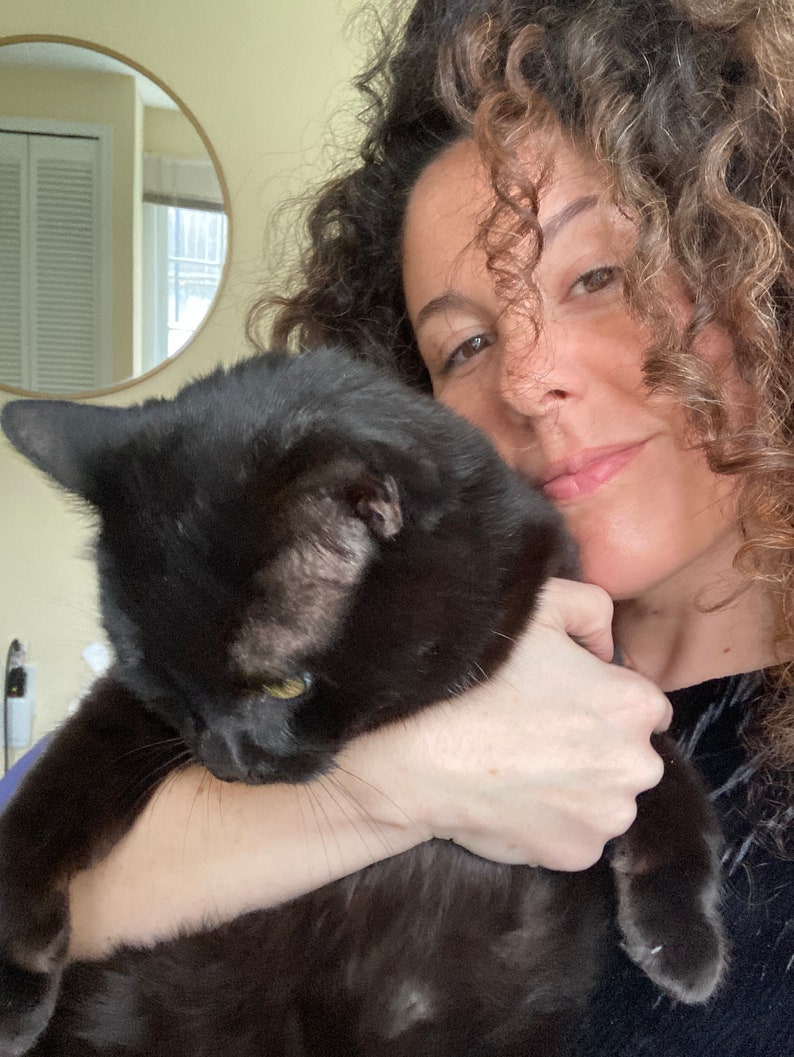 May include: A black cat with yellow eyes is being held. The cat's fur is shiny and smooth. The background includes a mirror and a person with curly hair.