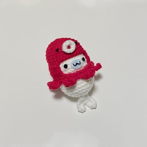 May include: A white crocheted plush toy wearing a red crocheted hat with a white circle on top. The toy has a white face with black eyes and a small blue mouth.