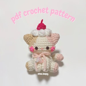 May include: A handmade crochet cat with a cupcake-shaped head. The cat is primarily cream-colored with pink and tan accents. It has pink star-shaped embellishments on its cheeks and a pink scarf. The text "pdf crochet pattern" is above the cat.