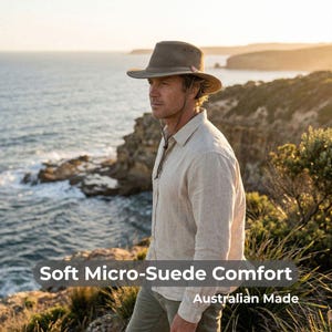 May include: A brown micro-suede hat with a chin strap, worn by a person. The image includes the text "Soft Micro-Suede Comfort" and "Australian Made". The background shows a coastal landscape with the sea and cliffs.