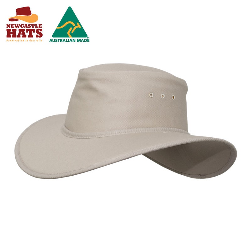 Op de afbeelding: Een beige hoed met een brede rand, een platte kroon en drie metalen ventilatiegaatjes aan de zijkant. De hoed toont de tekst NEWCASTLE HATS en AUSTRALIAN MADE met een kangoeroelogo in de linkerbovenhoek van de afbeelding.