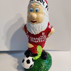 May include: A red and white gnome figurine wearing a football shirt with the text "Liverpool FC" and "Standard Chartered" on the front. The gnome is holding a black and white football and standing on a green base.