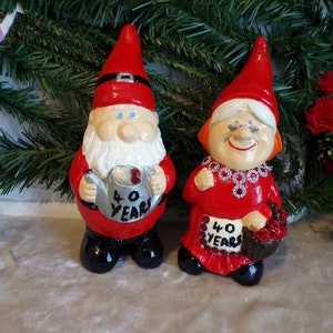 May include: Two red and white ceramic gnomes, one holding a watering can with the text "40 Years" and the other holding a basket with the same text. The gnomes are standing on a white surface.
