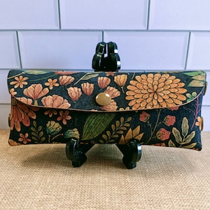 May include: A black fabric pencil case with a floral print in shades of orange, yellow, and green. The case has a gold button closure.