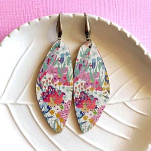 Pode incluir: Um par de brincos com padrão floral sobre fundo branco com flores rosa, azuis e amarelas. Os brincos são feitos de madeira e têm um gancho prateado.