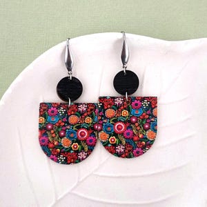 Könnte beinhalten: Ein Paar Ohrringe mit einem schwarzen Kreis und einem halbkreisförmigen Blumendesign. Das Blumendesign ist bunt mit rosa, roten, orangefarbenen, gelben, blauen und grünen Blumen auf schwarzem Hintergrund.