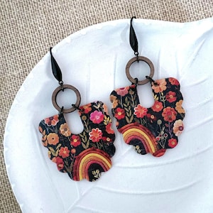 Könnte beinhalten: Ein Paar schwarze und braune Blumenohrringe mit einem Regenbogenmuster. Die Ohrringe sind aus Holz und haben einen gewellten Rand.