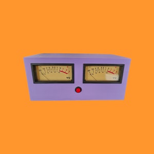 Op de afbeelding: Een lila-kleurige audio VU-meter met twee analoge displays. Elke display heeft een zwart frame, een gele wijzerplaat en een naald. Een rode knop is in het midden eronder. De achtergrond is oranje.