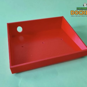 May include: A red rectangular tray with a hole and three smaller holes. The tray has a sloped side and the text "DOCKICON" is visible in the upper right corner. The tray is designed for a remote docking station.