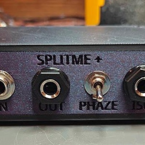 May include: A black box with the text "SPLITIME+" on the front. It has four jacks, two on the left and two on the right, and a toggle switch in the center labeled "PHASE".