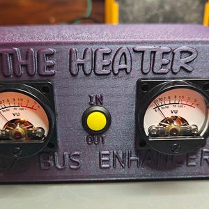 Op de afbeelding: Een paarse doos met de tekst "THE HEATER" en "MIX BUS ENHANCER" aan de voorkant. De doos heeft twee VU-meters met zwarte wijzerplaten en rode naalden, een gele knop met het label "IN/OUT" en een blauwe LED-lamp.