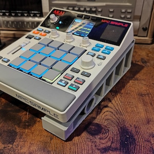 May include: A white and gray AKAI MPC SAMPLE music production center with blue illuminated pads and a screen. The device has various knobs and buttons for controlling sound. The unit is on a gray stand, with the words "MPC SAMPLE" printed on the front.