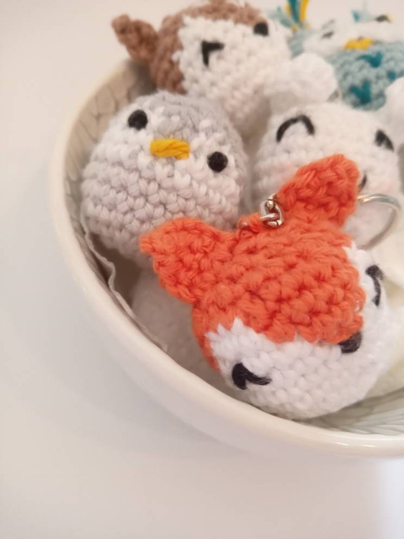 May include: A collection of crocheted animal keychains in a white bowl. The keychains include a penguin, a fox, a polar bear, and a blue and white animal.