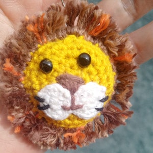 May include: A crocheted lion keychain with a yellow face, brown mane, and black eyes. The lion has a white nose and mouth.