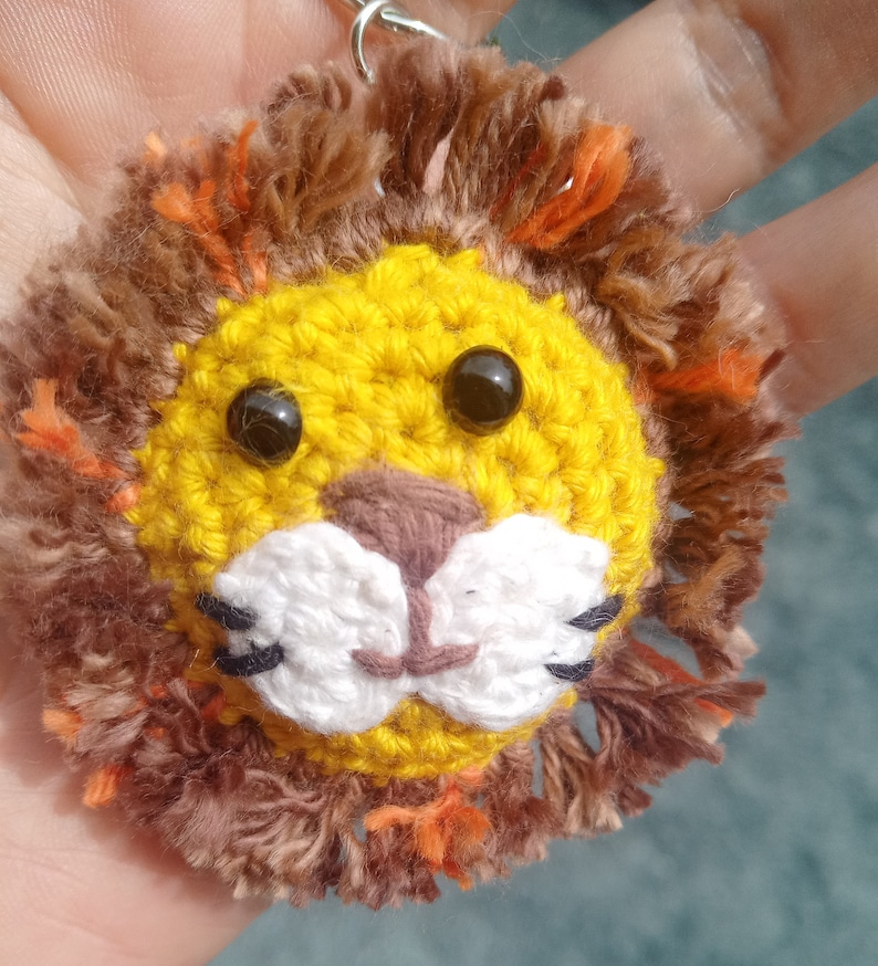 May include: A crocheted lion keychain with a yellow face, brown mane, and black eyes. The lion has a white nose and mouth.