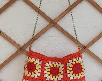 May include: An orange crochet shoulder bag with a chain strap. The bag features a pattern of red, yellow, and white floral squares. The bag is hanging against a wooden trellis.