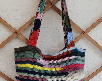 May include: A colorful, handmade crochet tote bag with a patchwork design. The bag features a variety of colors including red, blue, green, and yellow. The straps are made of a patterned fabric. The bag is displayed against a wooden lattice.