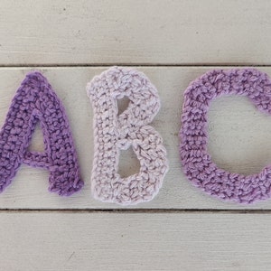 Könnte beinhalten: Häkelbuchstaben A, B und C in Violett- und Weißtönen. Die Buchstaben sind in einer Reihe auf einer weißen Holzoberfläche angeordnet.