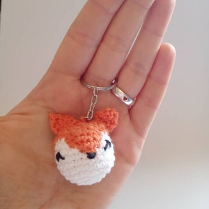 May include: A crocheted fox keychain with orange fur, white face, and black eyes. The keychain is attached to a silver key ring.